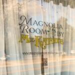 A window with white curtains behind it has a sticker in the front reading, "Magnolia Room by Miz Kathi's. www.magnoliaroom.mizkathys.com." In the reflection is a street with full parking lot on the other side.