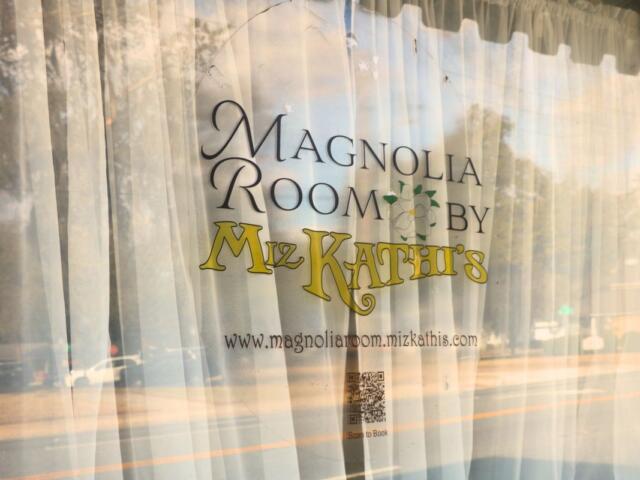 A window with white curtains behind it has a sticker in the front reading, "Magnolia Room by Miz Kathi's. www.magnoliaroom.mizkathys.com." In the reflection is a street with full parking lot on the other side.