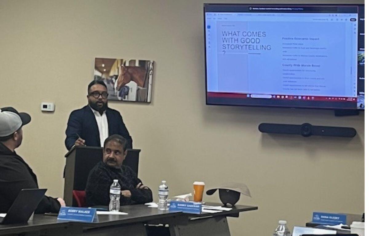 A man sitting down on the extreme left, with a table with bottles, paper name placards with a man siting in front of a podium, with another man standing behind the podium, and behind him on the wall is a picture of a horse, and to the right against a beige wall is a large screen displaying a slide.