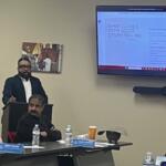 A man sitting down on the extreme left, with a table with bottles, paper name placards with a man siting in front of a podium, with another man standing behind the podium, and behind him on the wall is a picture of a horse, and to the right against a beige wall is a large screen displaying a slide.