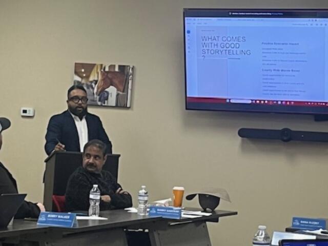 A man sitting down on the extreme left, with a table with bottles, paper name placards with a man siting in front of a podium, with another man standing behind the podium, and behind him on the wall is a picture of a horse, and to the right against a beige wall is a large screen displaying a slide.