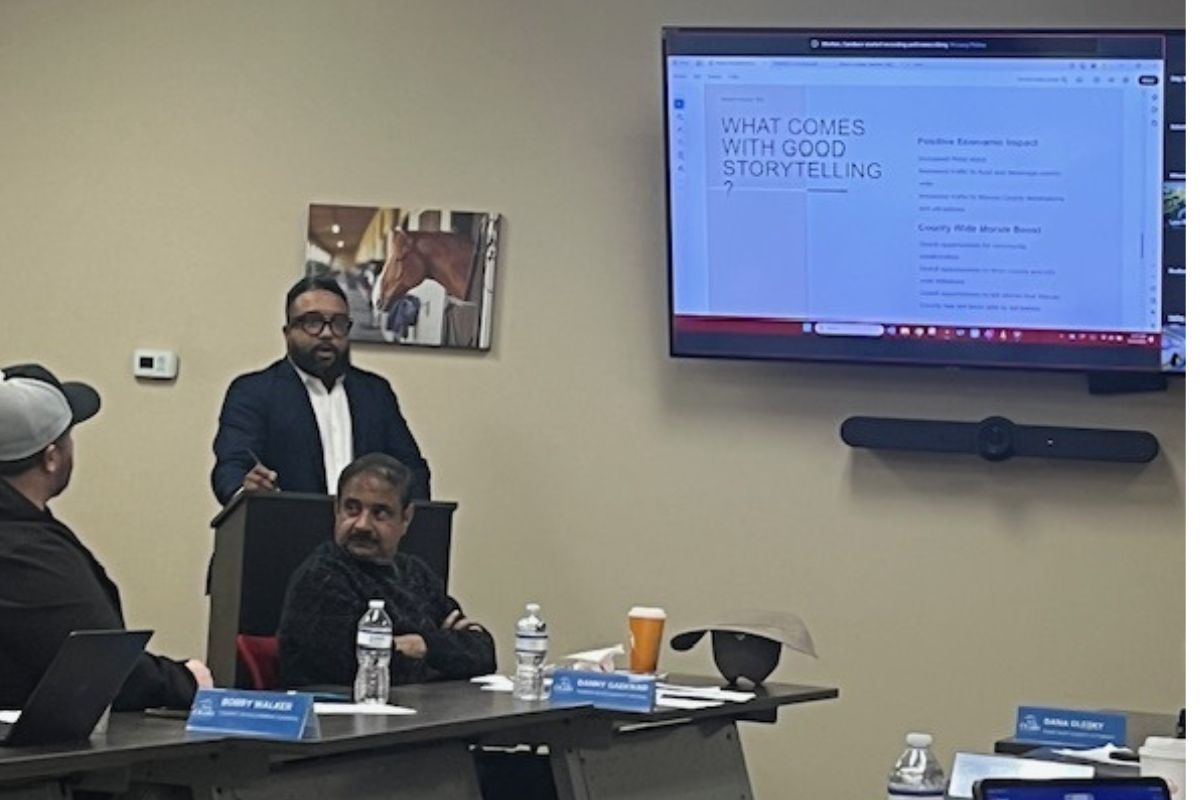 A man sitting down on the extreme left, with a table with bottles, paper name placards with a man siting in front of a podium, with another man standing behind the podium, and behind him on the wall is a picture of a horse, and to the right against a beige wall is a large screen displaying a slide.