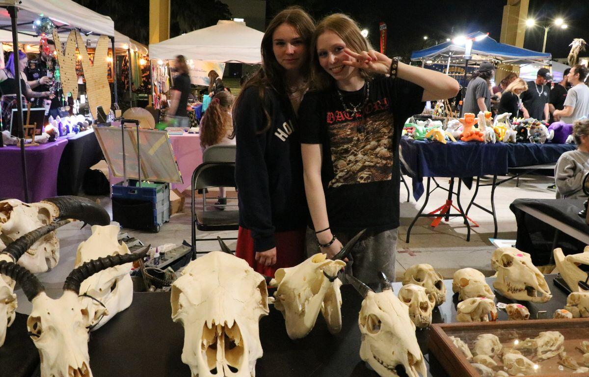 Two girls stand behind a table with a black tablecloth with animal skulls scattered across it. Some smaller skulls are in a wooden display case next to the large skulls.