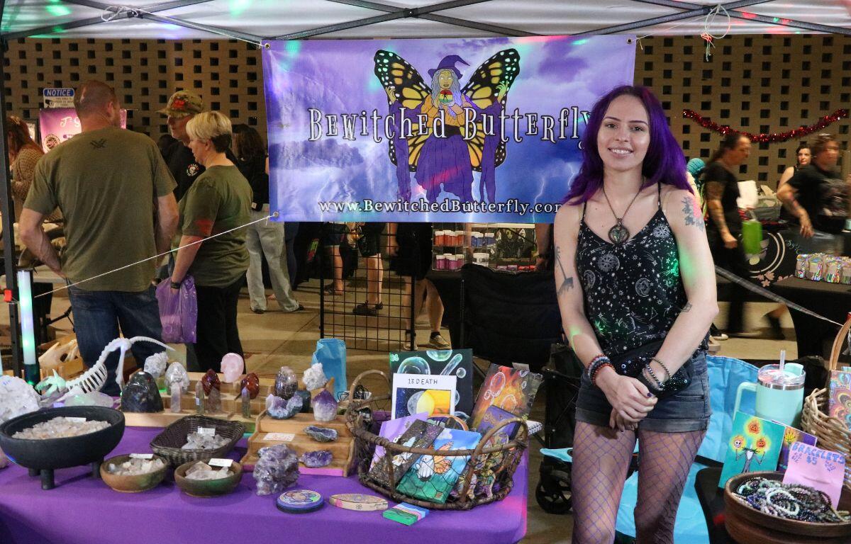 A woman stands next to a table with a basket full of canvases and crystals displayed on shelves and in bowls. behind her is a sign that reads, "Bewitched Butterfly."