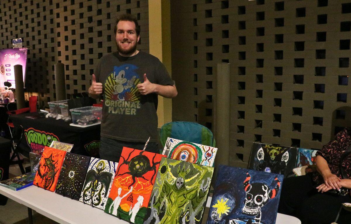 A man stands behind a table with multiple horror-themed paintings lined up on a white table.