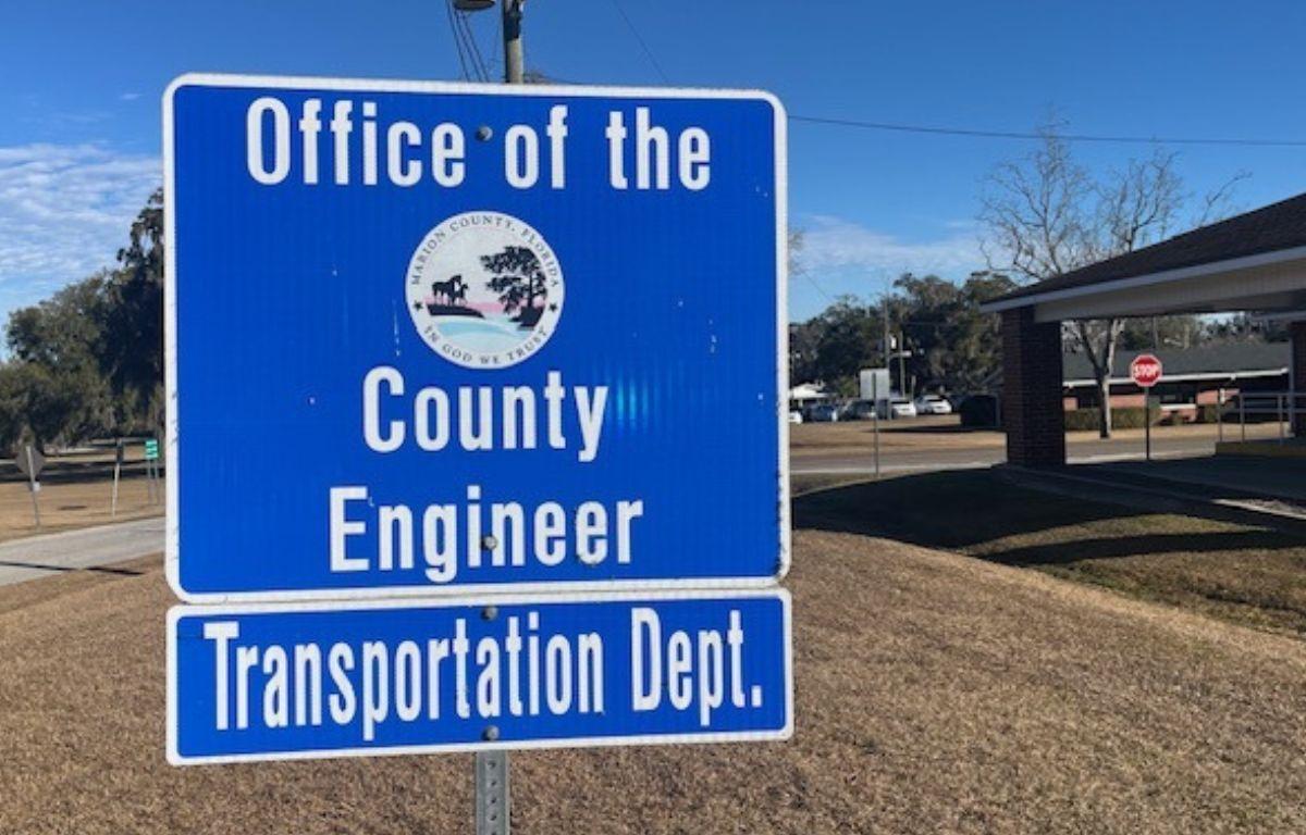 A blue sign with white lettering, and a government entity insignia against the blue background. In the foreground and background, a lawn, and to the right trees and a building.