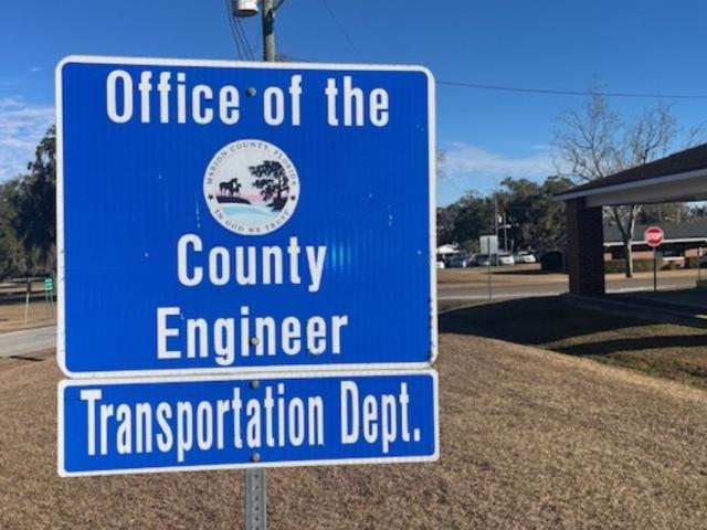 A blue sign with white lettering, and a government entity insignia against the blue background. In the foreground and background, a lawn, and to the right trees and a building.
