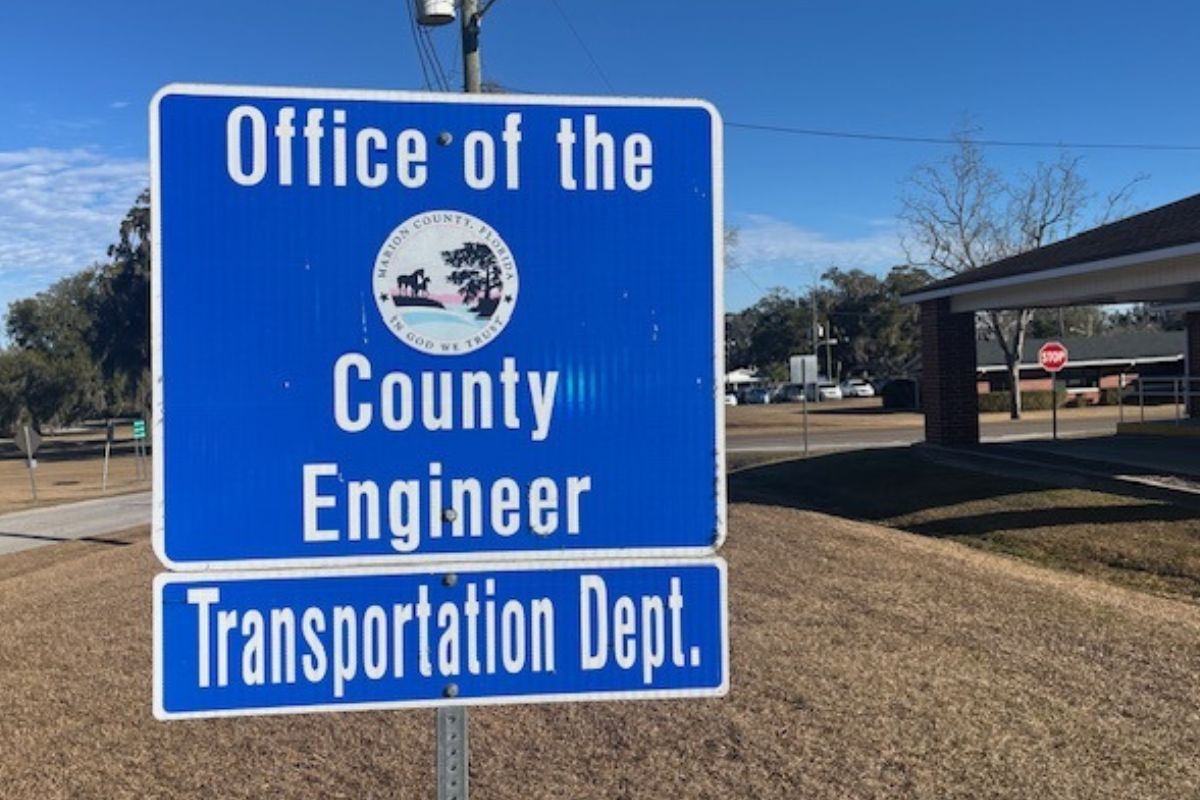 A blue sign with white lettering, and a government entity insignia against the blue background. In the foreground and background, a lawn, and to the right trees and a building.