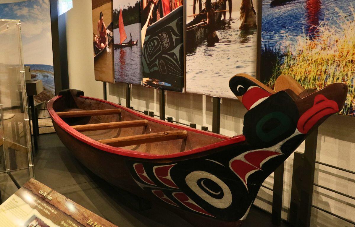 A dugout sits against a wall in a museum with painted designs. The background feature pictures on the walls of people rowing in dugouts.