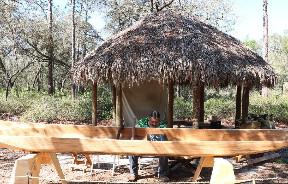 A man sits in front of a chikee hut with a wooden canoe in front of him. A rope is strung along in front of it to keep onlookers from touching it.