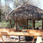 A man sits in front of a chikee hut with a wooden canoe in front of him. A rope is strung along in front of it to keep onlookers from touching it.