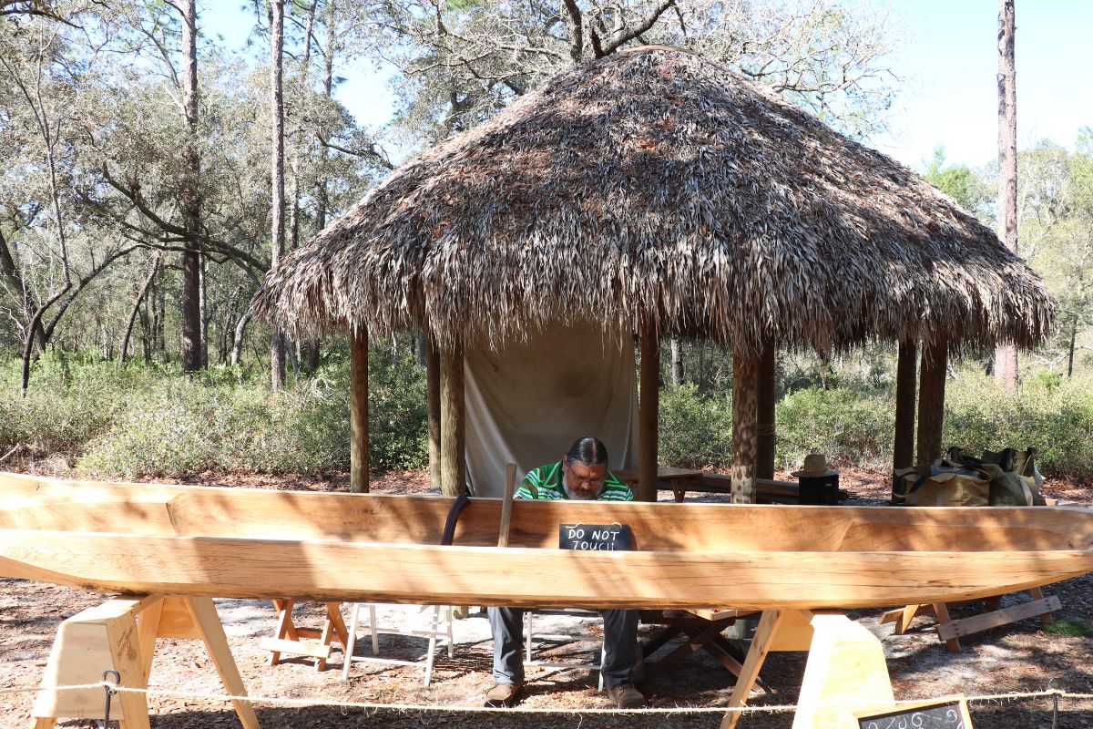 A man sits in front of a chikee hut with a wooden canoe in front of him. A rope is strung along in front of it to keep onlookers from touching it.