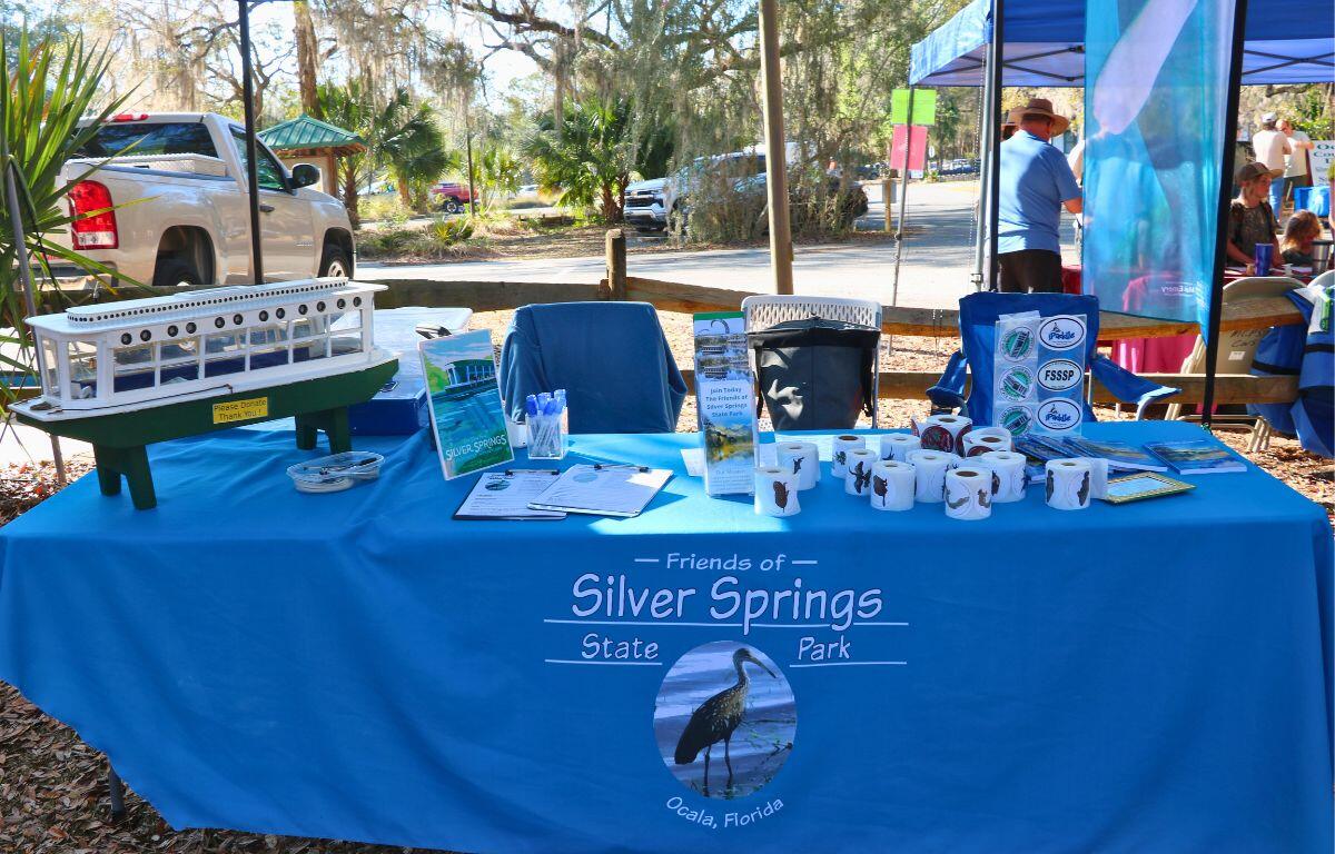 A vendor booth features a blue tablecloth that reads, "Friends of Silver Springs State Park." On the table are promotional and informational items about Silver Springs State Park.