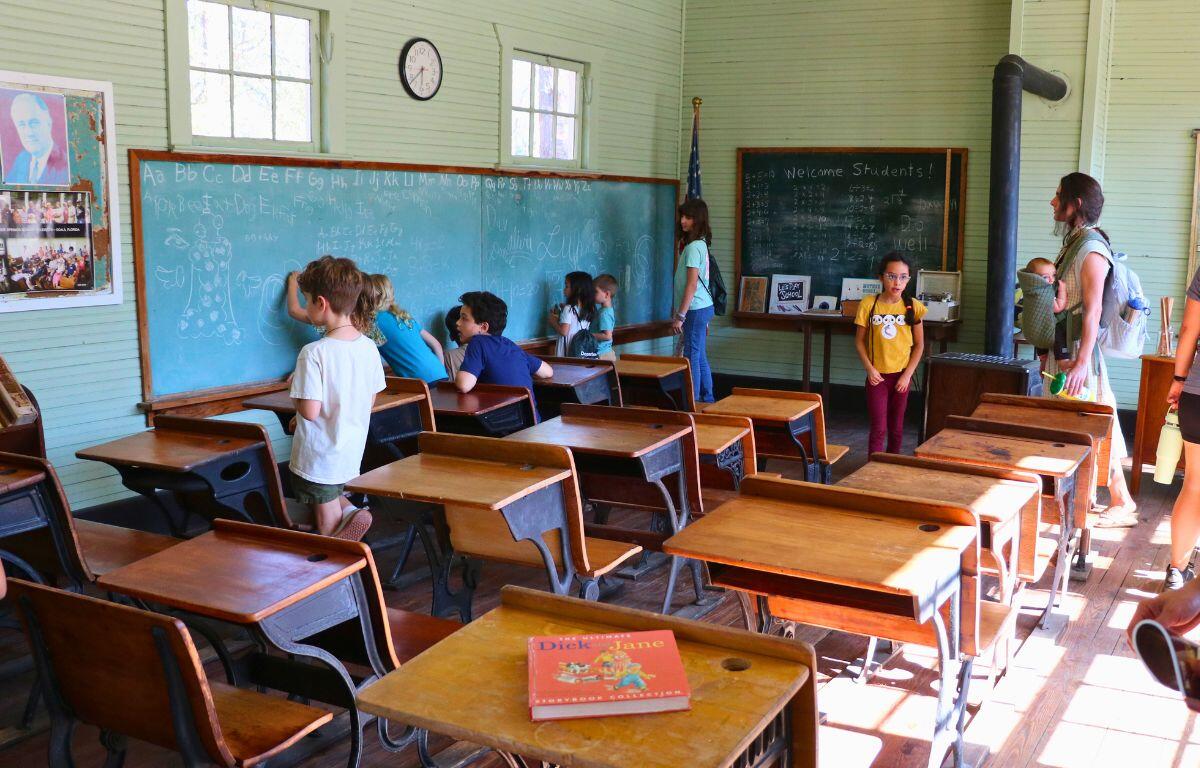 Kids are gathered in front of a chalkboard in a historic schoolhouse, drawing and writing with the chalk.