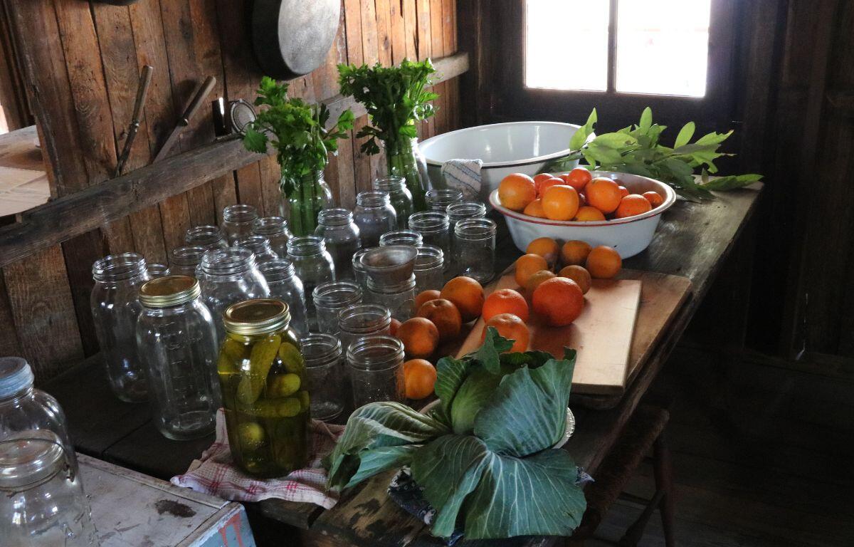 Oranges are scattered across a wooden table with more inside a large, white bowl. Vegetables are displayed in glass jars.