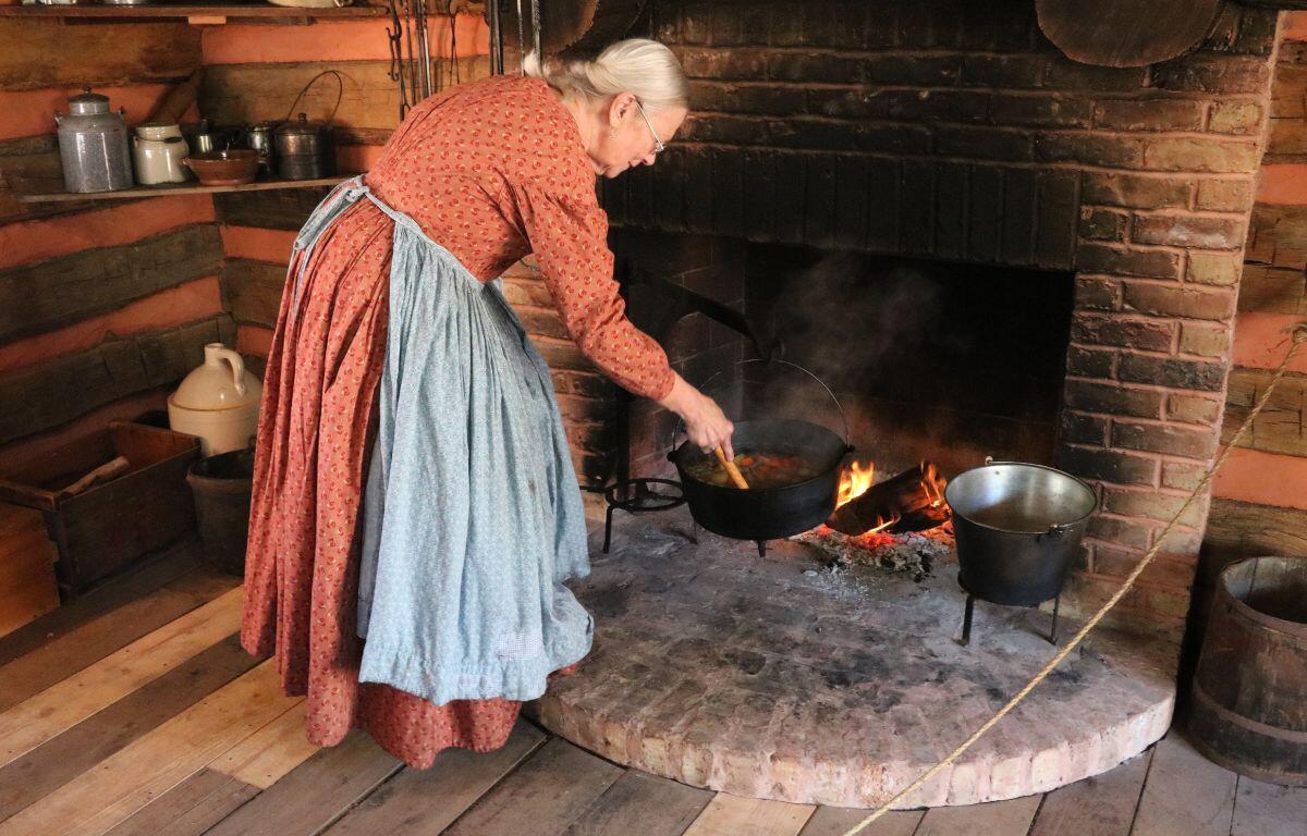A woman in 1800s-themed attire stirs stew over a fire.
