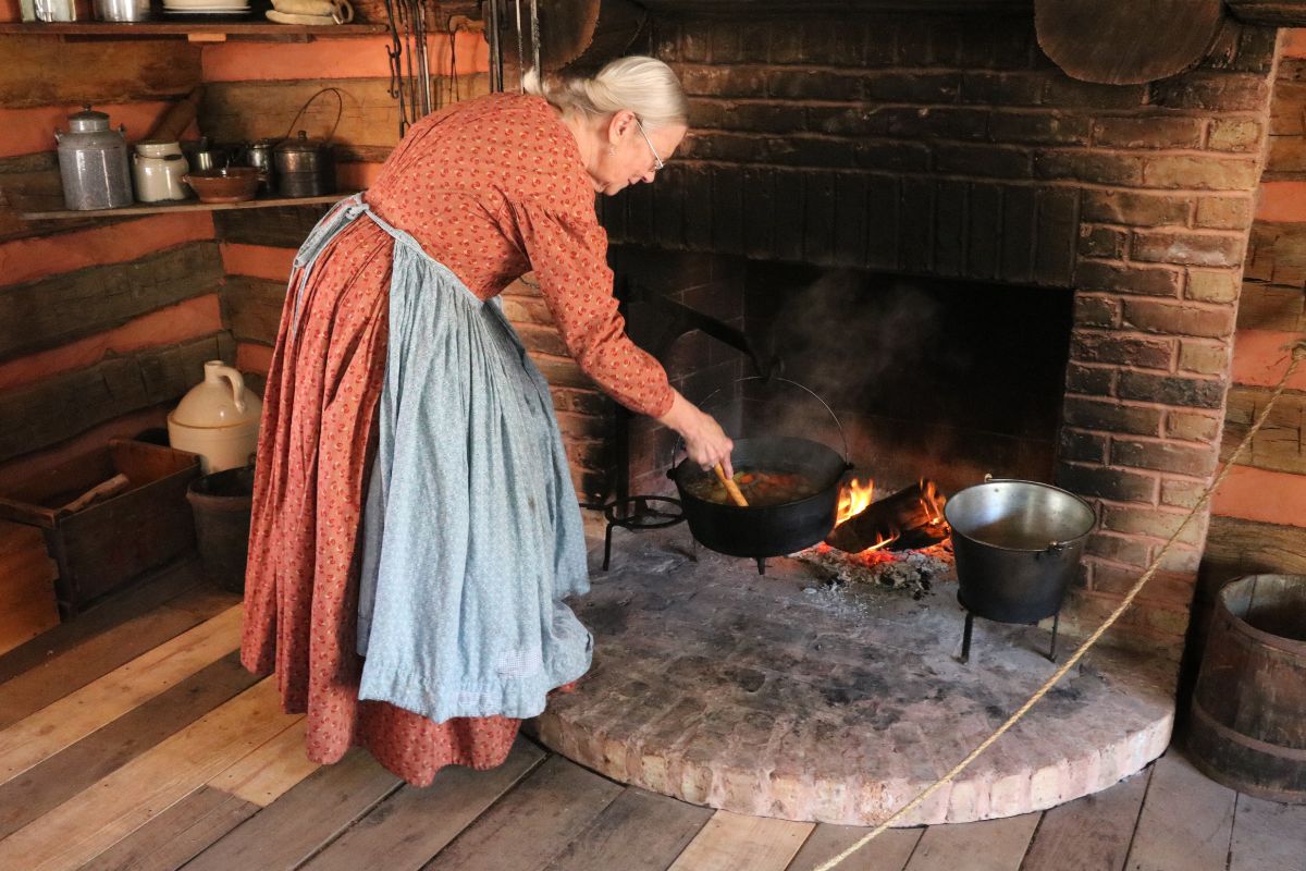A woman in 1800s-themed attire stirs stew over a fire.