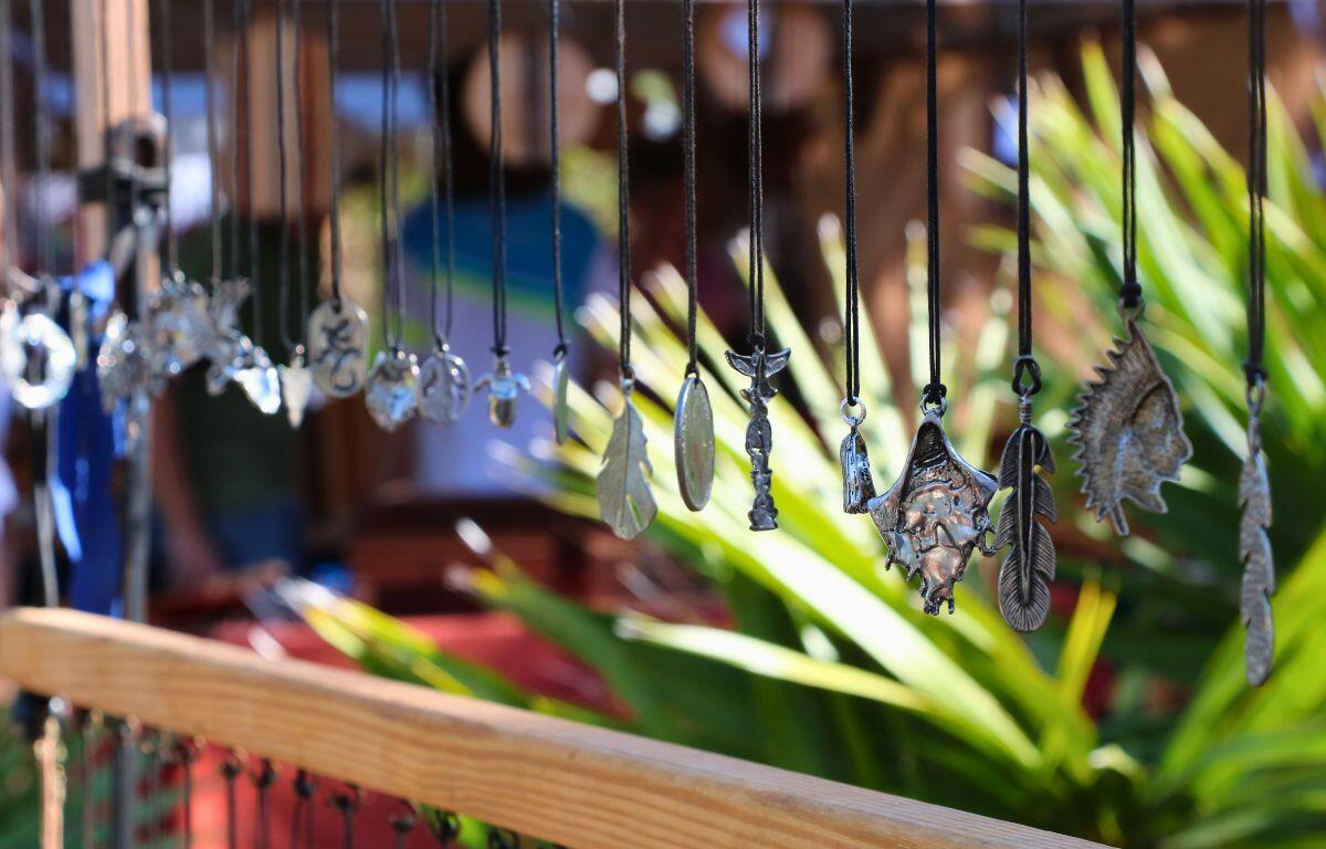 Metal necklaces with varying shapes and designs hang from a wood shelf with trees and a market in the background.