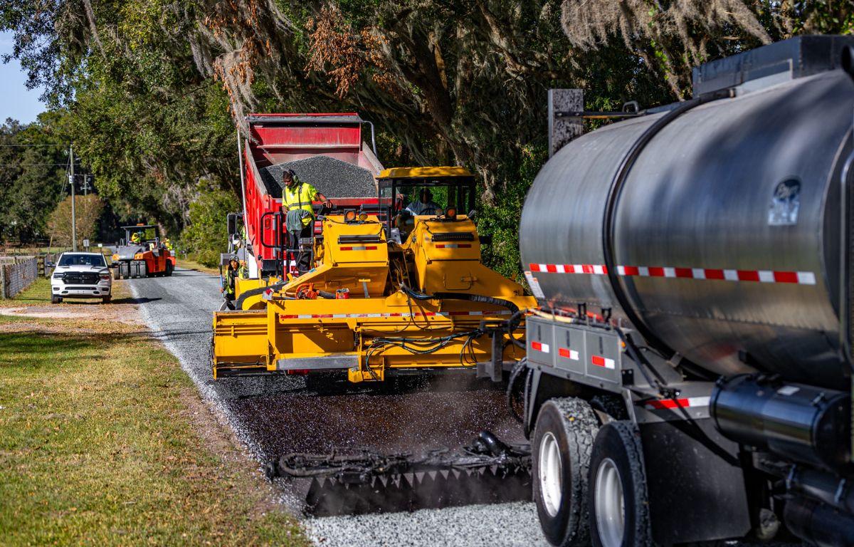 On the left a grassy median, in the background, live oak trees with Spanish moss, in the foreground a tanker, and in the middle a paver laying down asphalt and resurfacing the road.