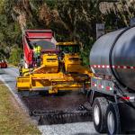 On the left a grassy median, in the background, live oak trees with Spanish moss, in the foreground a tanker, and in the middle a paver laying down asphalt and resurfacing the road.