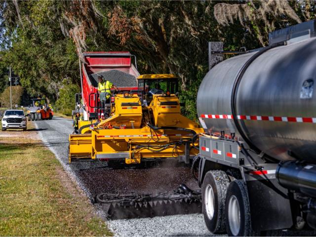 On the left a grassy median, in the background, live oak trees with Spanish moss, in the foreground a tanker, and in the middle a paver laying down asphalt and resurfacing the road.
