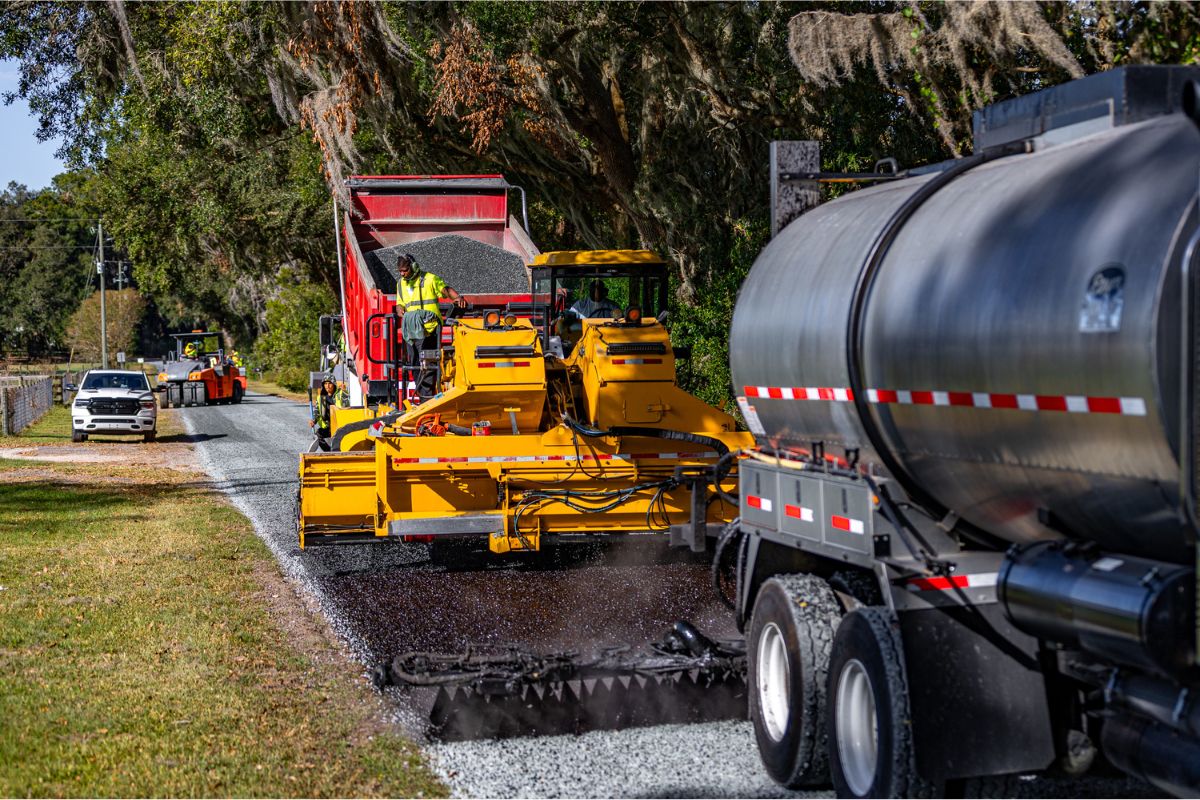 On the left a grassy median, in the background, live oak trees with Spanish moss, in the foreground a tanker, and in the middle a paver laying down asphalt and resurfacing the road.