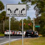 A road sign with arrows pointing in different directions, in the background, vehicles, waiting at a traffic light, trees to the left and right, with a street sign to the right.
