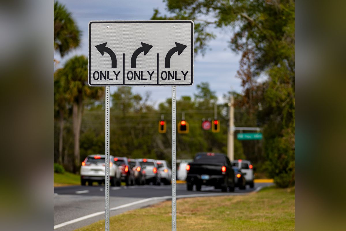A road sign with arrows pointing in different directions, in the background, vehicles, waiting at a traffic light, trees to the left and right, with a street sign to the right.