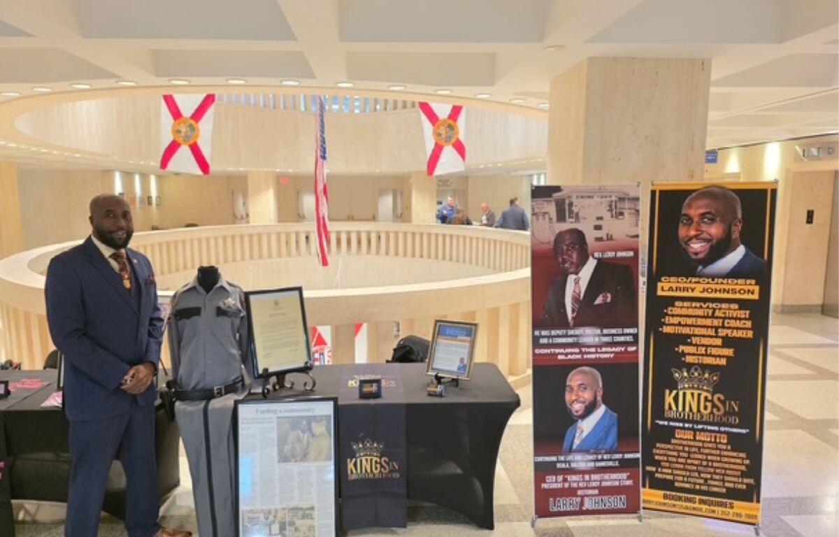 A man standing in front of a circular rotunda, with glass, and flags suspended from the ceiling in the background. The man is to the left of a number of items, including a uniform, framed documents on the floor and on a table to the right of a man, and picture placards to the extreme right.