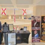 A man standing in front of a circular rotunda, with glass, and flags suspended from the ceiling in the background. The man is to the left of a number of items, including a uniform, framed documents on the floor and on a table to the right of a man, and picture placards to the extreme right.