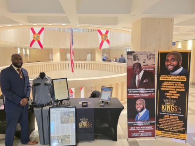 A man standing in front of a circular rotunda, with glass, and flags suspended from the ceiling in the background. The man is to the left of a number of items, including a uniform, framed documents on the floor and on a table to the right of a man, and picture placards to the extreme right.