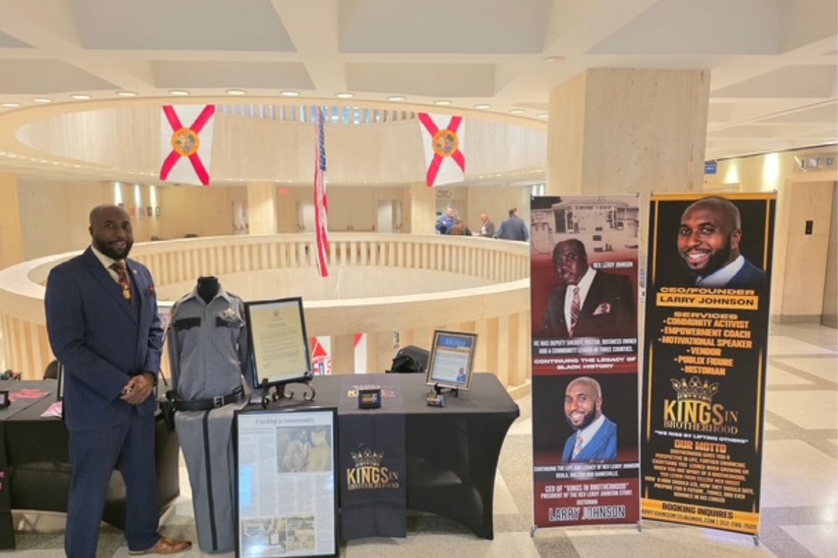A man standing in front of a circular rotunda, with glass, and flags suspended from the ceiling in the background. The man is to the left of a number of items, including a uniform, framed documents on the floor and on a table to the right of a man, and picture placards to the extreme right.