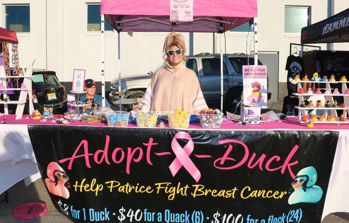 A woman stands behind a booth with a long table full of ducks and small accessories on top of a pink tablecloth. In front of the booth is a banner reading, "Adopt-A-Duck. Help Patrice Fight Breast Cancer."
