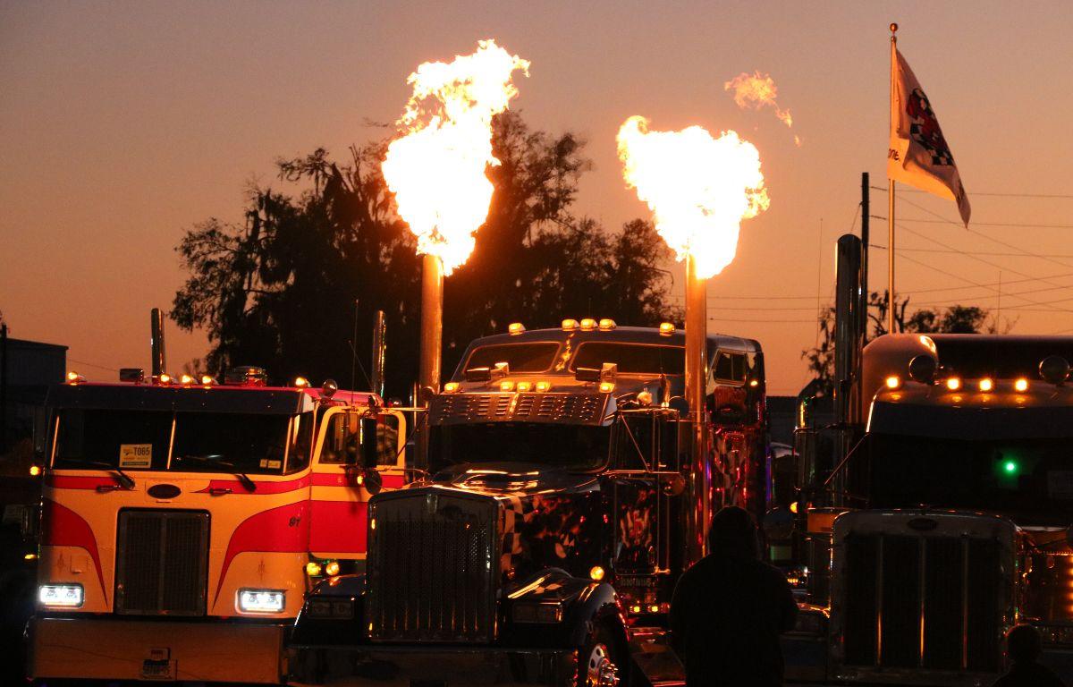 A semi-truck blows flames through its exhaust while other tucks around it are lit by the flames.