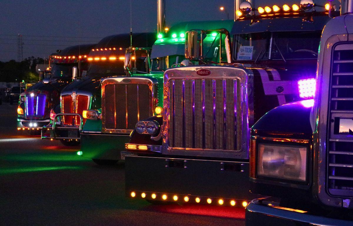Trucks are lined up in a parking lot with bright lights shining from them with a dark sky behind them.