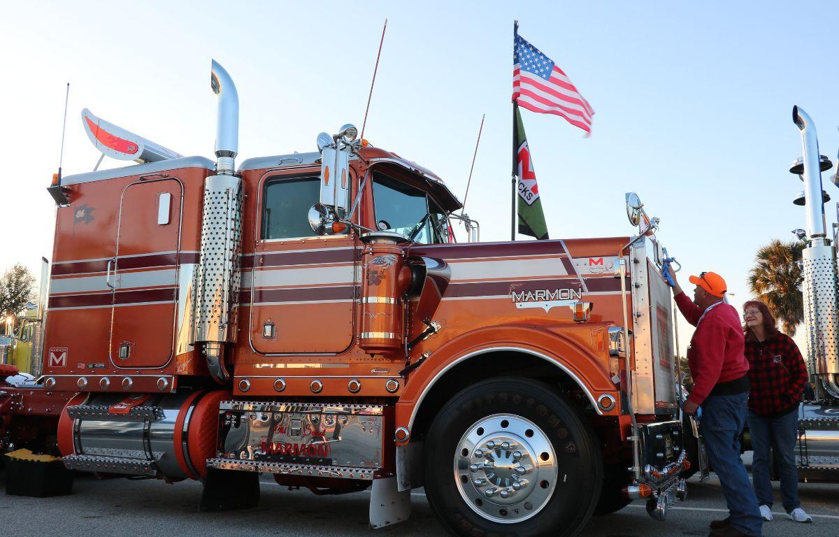 A large, orange truck with brown and white stripes sits in a parking lot while two people look at the grill. Flags are raised in the background.