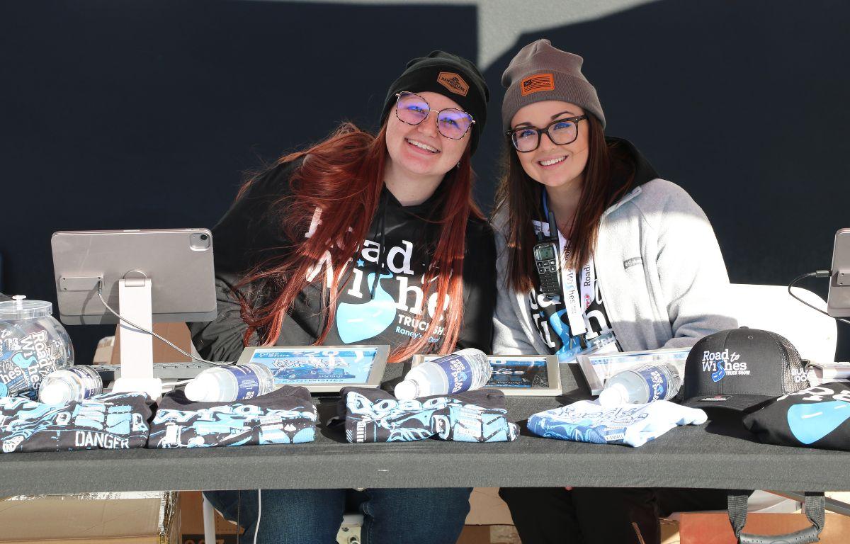 Two women sit behind a vendor booth with shirts, water, hats and information flyers.