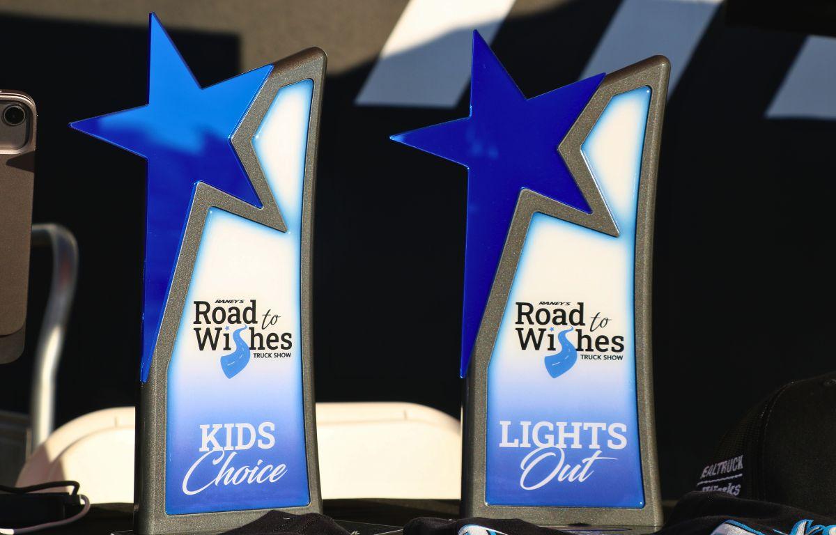 Two awards sit on a table with a black tablecloth with a black and white background. The awards read, "Road to Wishes Truck Show" with categories reading, "Kids Choice" and "Lights Out."
