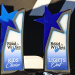 Two awards sit on a table with a black tablecloth with a black and white background. The awards read, "Road to Wishes Truck Show" with categories reading, "Kids Choice" and "Lights Out."