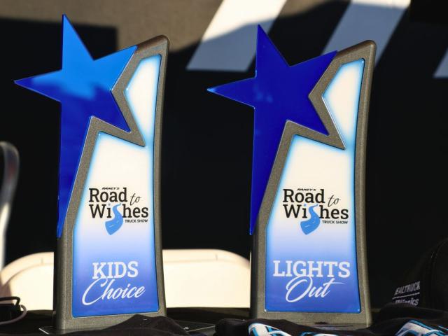 Two awards sit on a table with a black tablecloth with a black and white background. The awards read, "Road to Wishes Truck Show" with categories reading, "Kids Choice" and "Lights Out."