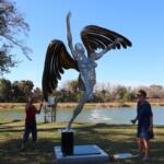 A large sculpture of a woman with wings making her way upward stands in a park with people and trees surrounding with a lake in the background.