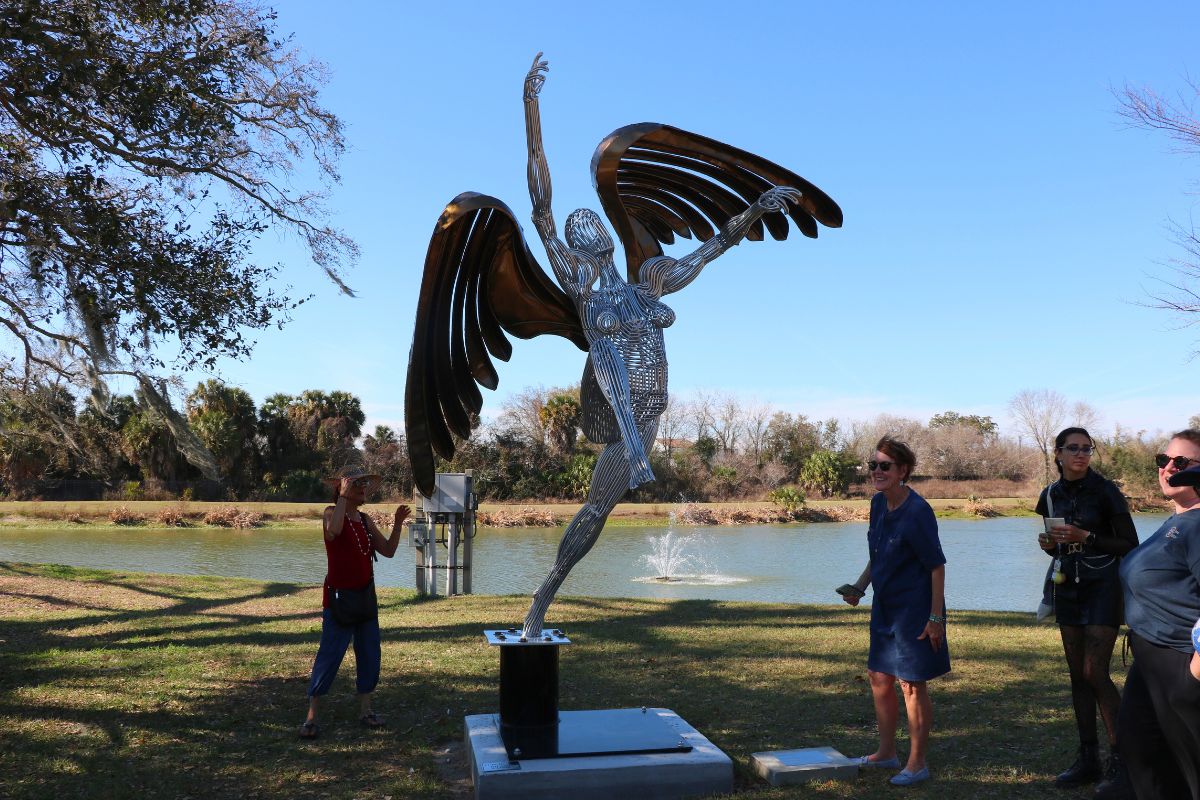 A large sculpture of a woman with wings making her way upward stands in a park with people and trees surrounding with a lake in the background.