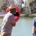 A man stands next to a sculpture, making a joke to people around him. The sculpture stands in front of a lake and features a large mouth.