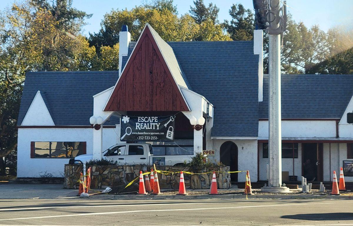Colored picture with a white historic building with a grey roof and wood accent. A sign is displayed, advertising "Escape Reality."