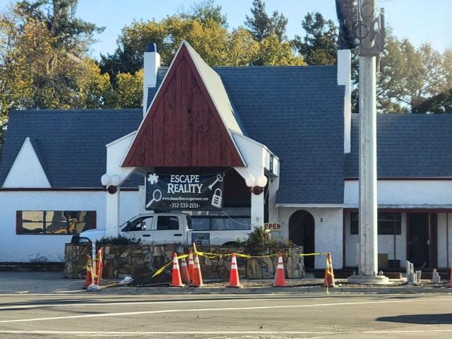 Colored picture with a white historic building with a grey roof and wood accent. A sign is displayed, advertising "Escape Reality."