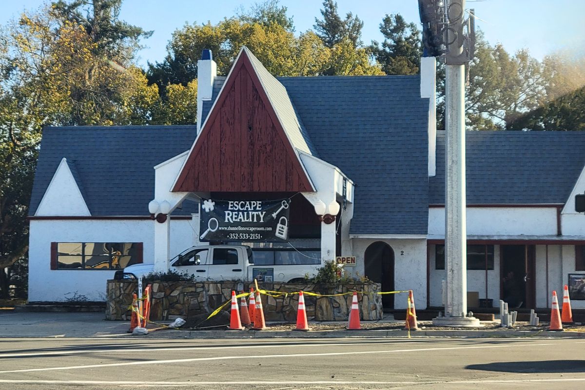 Colored picture with a white historic building with a grey roof and wood accent. A sign is displayed, advertising "Escape Reality."