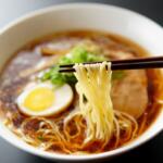A white bowl full of ramen noodles, boiled egg and seaweed and scallions in a broth with chopsticks.