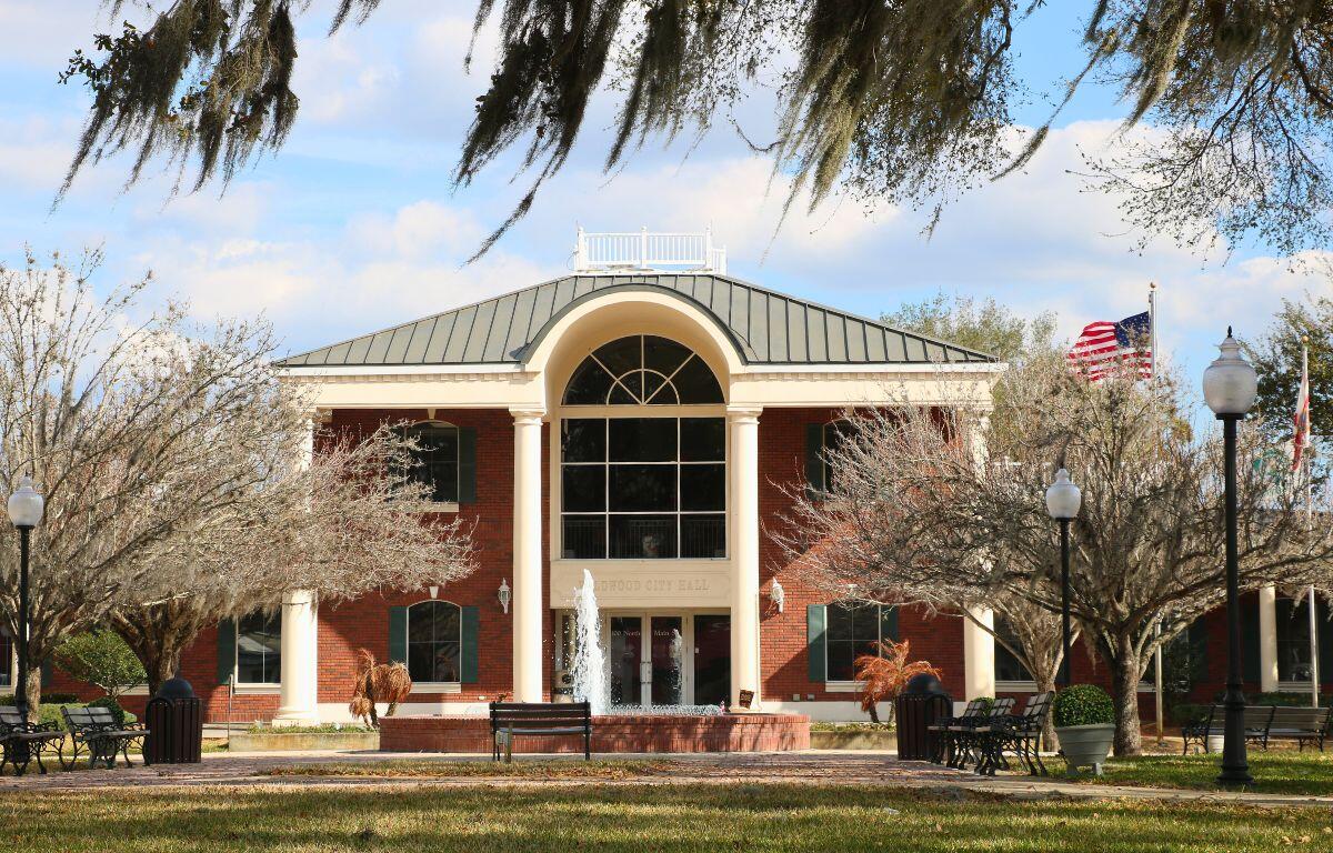 A large brick building with white pillars and large, glass windows stand in the middle of a busy town. In front the building is a fountain spouting water upward, benches, a flagpole, lamp posts, and trees.