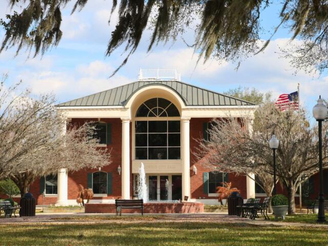 A large brick building with white pillars and large, glass windows stand in the middle of a busy town. In front the building is a fountain spouting water upward, benches, a flagpole, lamp posts, and trees.