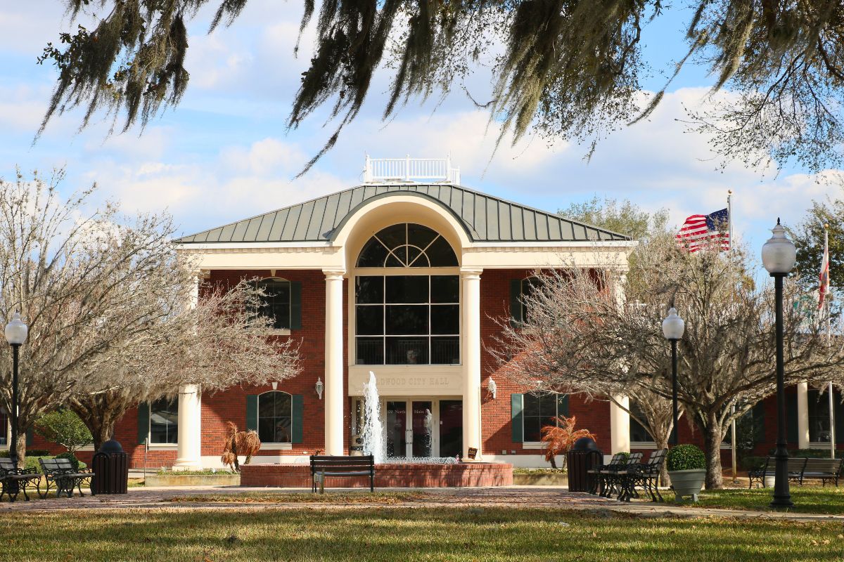 A large brick building with white pillars and large, glass windows stand in the middle of a busy town. In front the building is a fountain spouting water upward, benches, a flagpole, lamp posts, and trees.